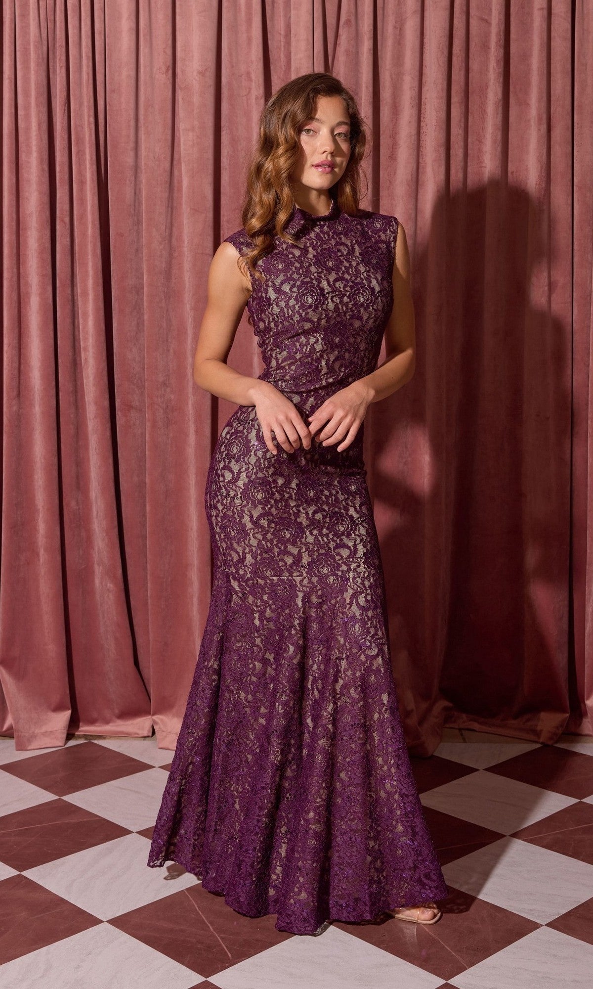 A woman stands on a checkered floor in front of mauve curtains, wearing the Eureka 2061 Long Lace Formal Dress in plum/gold—a sleeveless, floor-length lace evening gown.